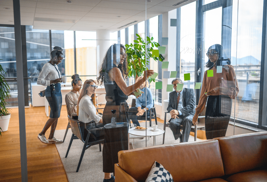 Diverse team in a modern office engaging in a collaborative workshop, with participants discussing ideas and placing sticky notes on glass—illustrating the reflective and strategic nature of team coaching compared to traditional team building.